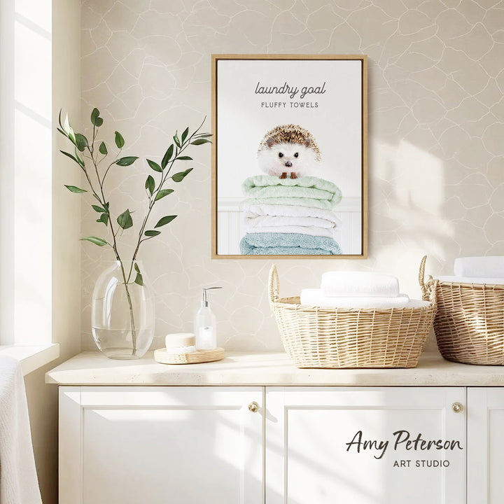 a framed poster of a hedgehog on a shelf, with a basket of towels below it. The poster has the text "laundry goal" and "fluffy towels" written on it. The shelf also has a vase with leaves and a bottle of soap.