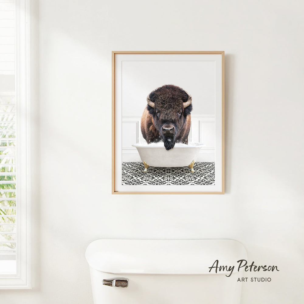 A framed image of a buffalo sitting in a bathtub, with a patterned tile floor beneath it, is hanging on a white wall.