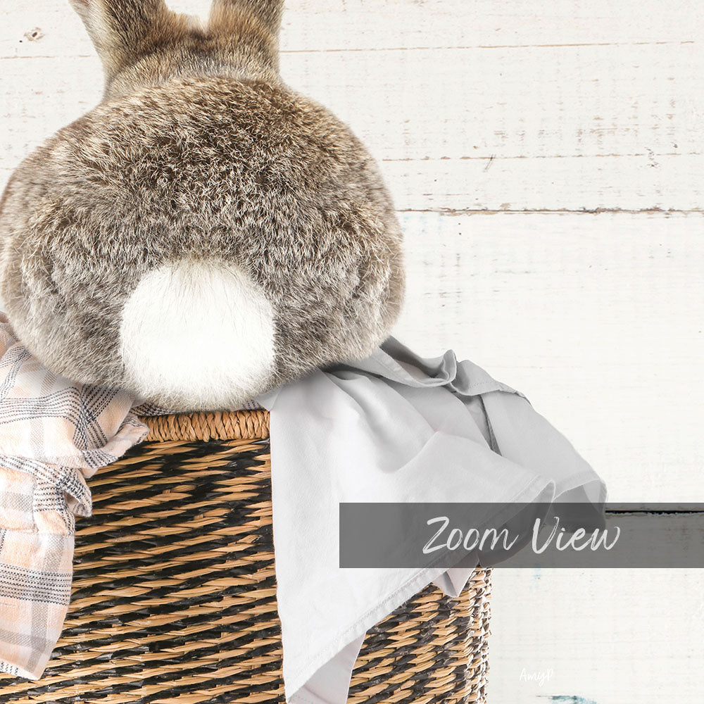 A fluffy gray rabbit sits in a woven basket, with a gray blanket and a white cloth nearby.