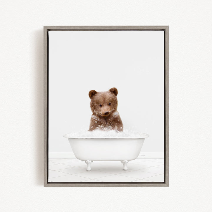 A cute brown bear sitting in a white bathtub filled with bubbles, looking directly at the camera.