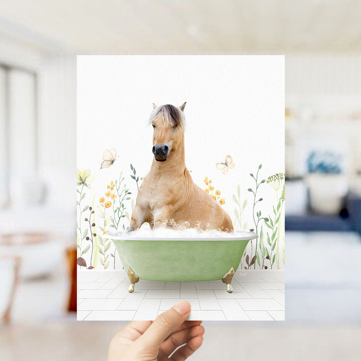 A hand holds up a framed image of a horse sitting in a green bathtub, surrounded by flowers and butterflies.