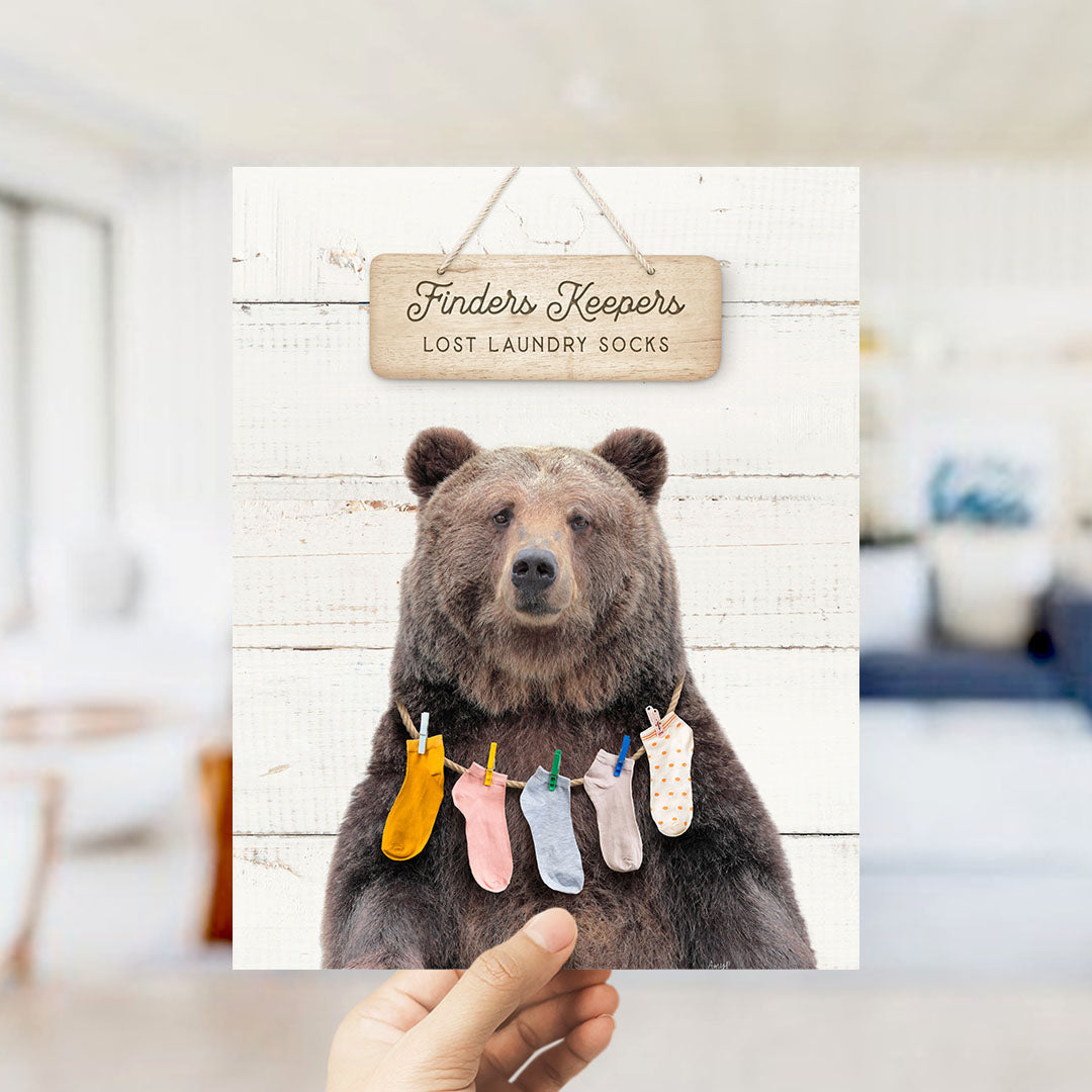 A hand holding a sign that says "Finders Keepers" above a bear wearing socks.