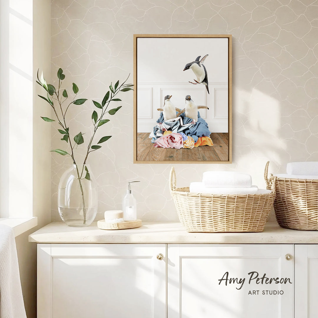 a bathroom setting with a framed picture of penguins on the wall, a vase with green leaves, and baskets on a counter.
