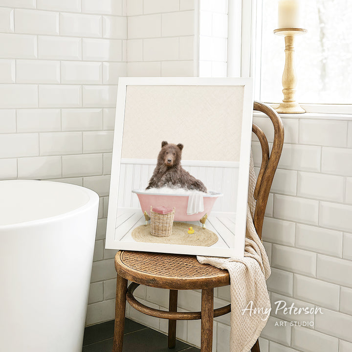 A framed image of a brown bear sitting in a pink bathtub, surrounded by a basket of bath toys, is displayed on a wooden chair in a bathroom setting.
