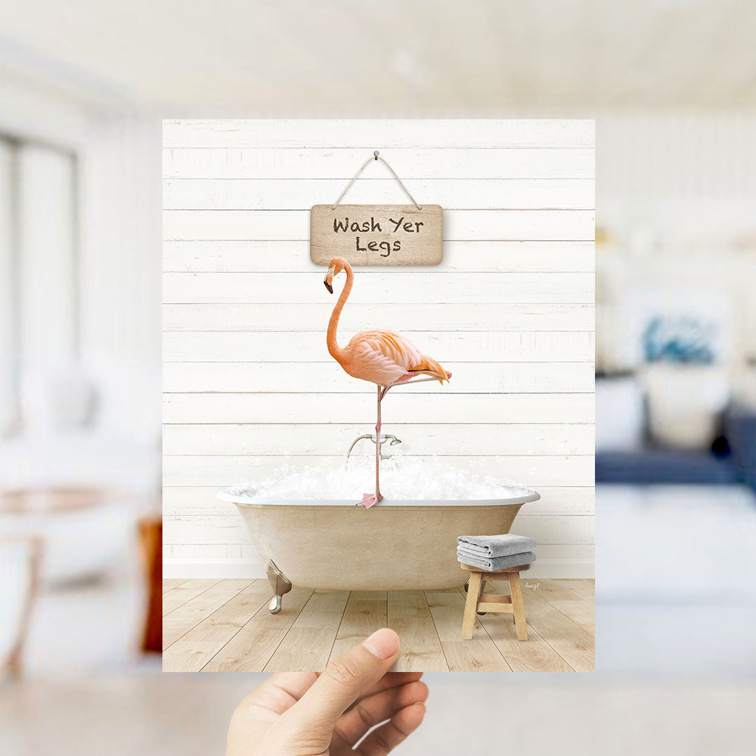 A hand holds up a framed image of a pink flamingo standing in a bathtub filled with bubbles, with a sign above it that says "Wash Yer Legs".