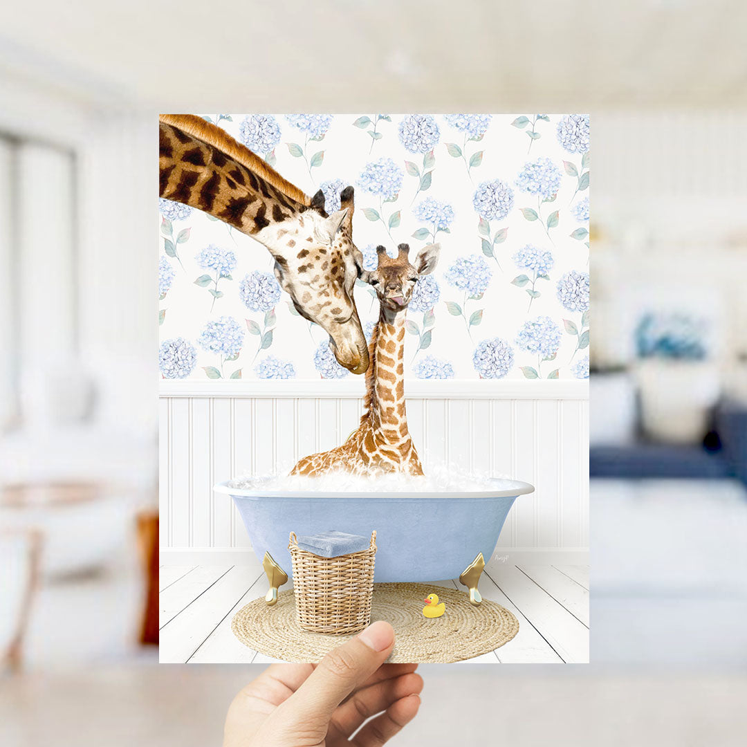 A hand holds up a framed photograph of two giraffes in a bathtub, with one giraffe sitting in the tub and the other standing nearby.