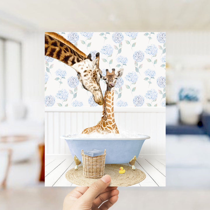 A hand holds up a framed photograph of two giraffes in a bathtub, with one giraffe sitting in the tub and the other standing nearby.