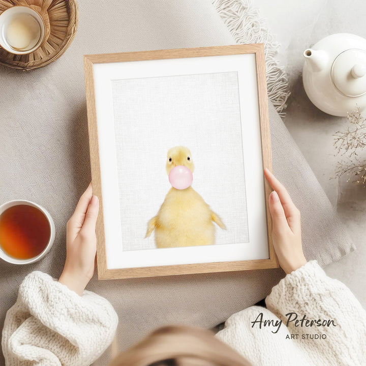 A person is holding a framed picture of a yellow duck blowing a bubble with a pink bubblegum bubble.