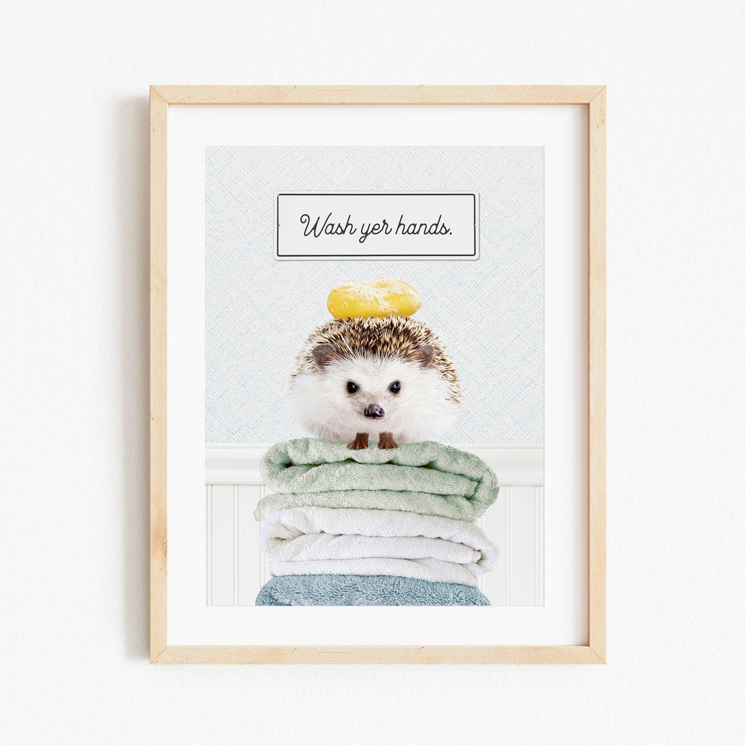 A framed image of a hedgehog sitting atop a stack of folded towels, with the text "Wash your hands" written above it.