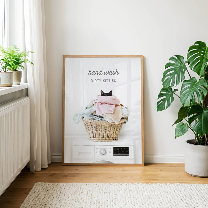 A framed poster on a wall, featuring a cat sitting in a basket filled with laundry, with the text "hand wash dirty kitties" and a washing machine icon.