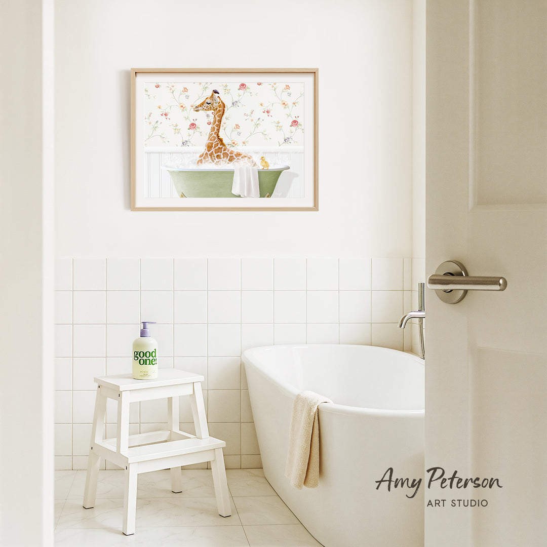 A framed painting of a giraffe sitting in a bathtub is hanging on a white tiled wall above a white bathtub in a bathroom.