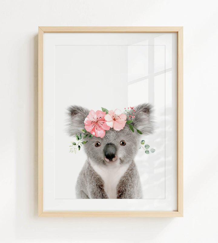 A framed image of a koala bear wearing a floral crown of pink flowers and greenery.