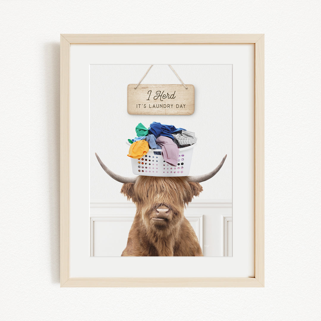 A framed image of a Highland cow with a basket of laundry on its head, accompanied by a wooden sign that reads "I Harded it's laundry day".