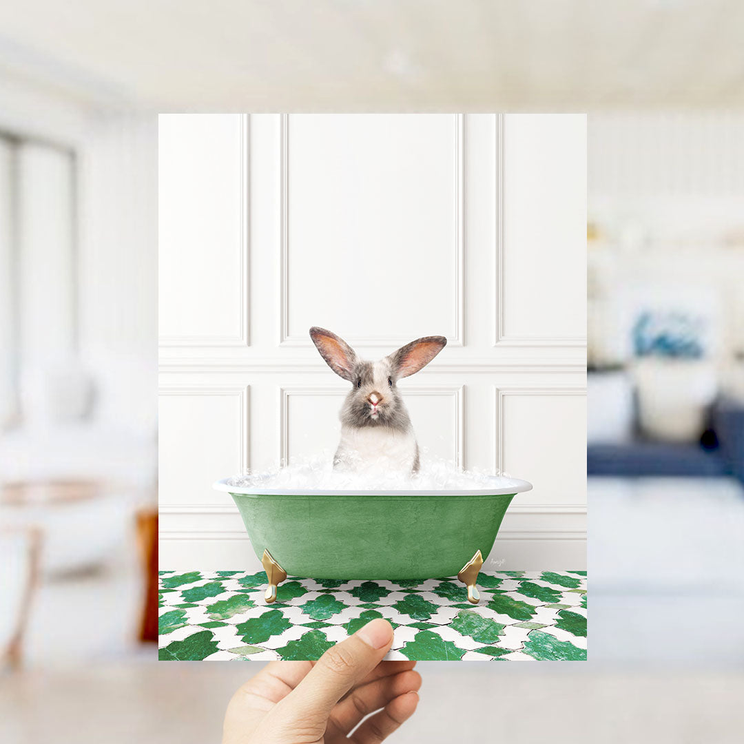 A hand holds up a framed image of a rabbit sitting in a green bathtub filled with bubbles, with a white wall and green and white patterned floor in the background.