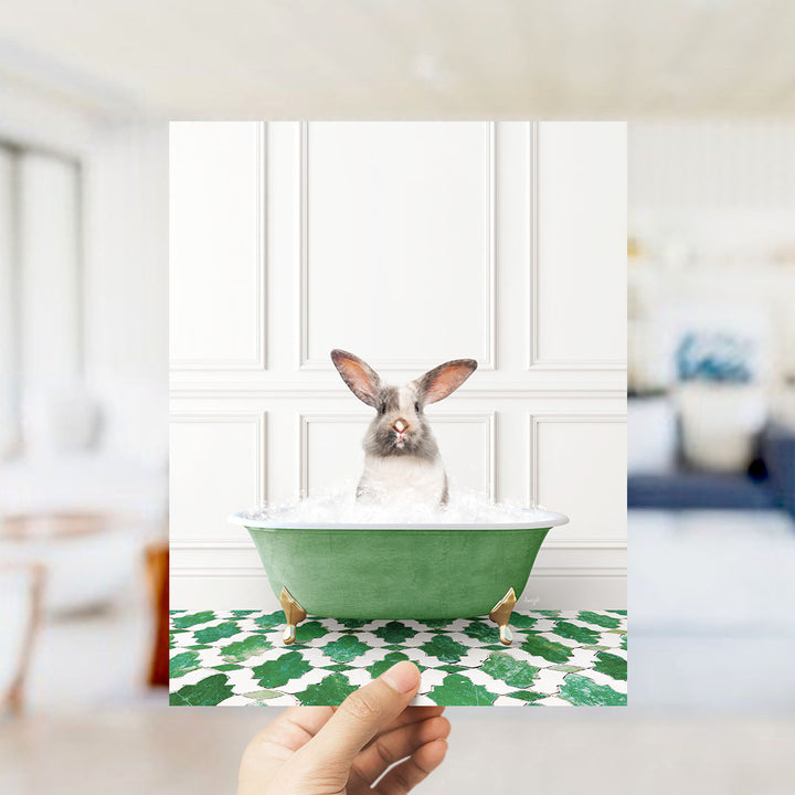 A hand holds up a framed image of a rabbit sitting in a green bathtub filled with bubbles, with a white wall and green and white patterned floor in the background.