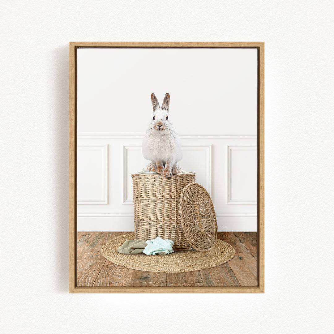 A white rabbit sits atop a woven basket on a wooden floor, with a green blanket and a woven basket nearby.