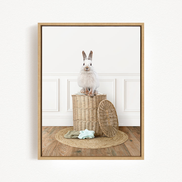A white rabbit sits atop a woven basket on a wooden floor, with a green blanket and a woven basket nearby.