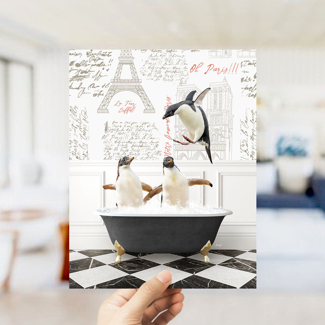 A hand is holding up a framed image depicting three penguins sitting in a bathtub, with the Eiffel Tower and Paris in the background.