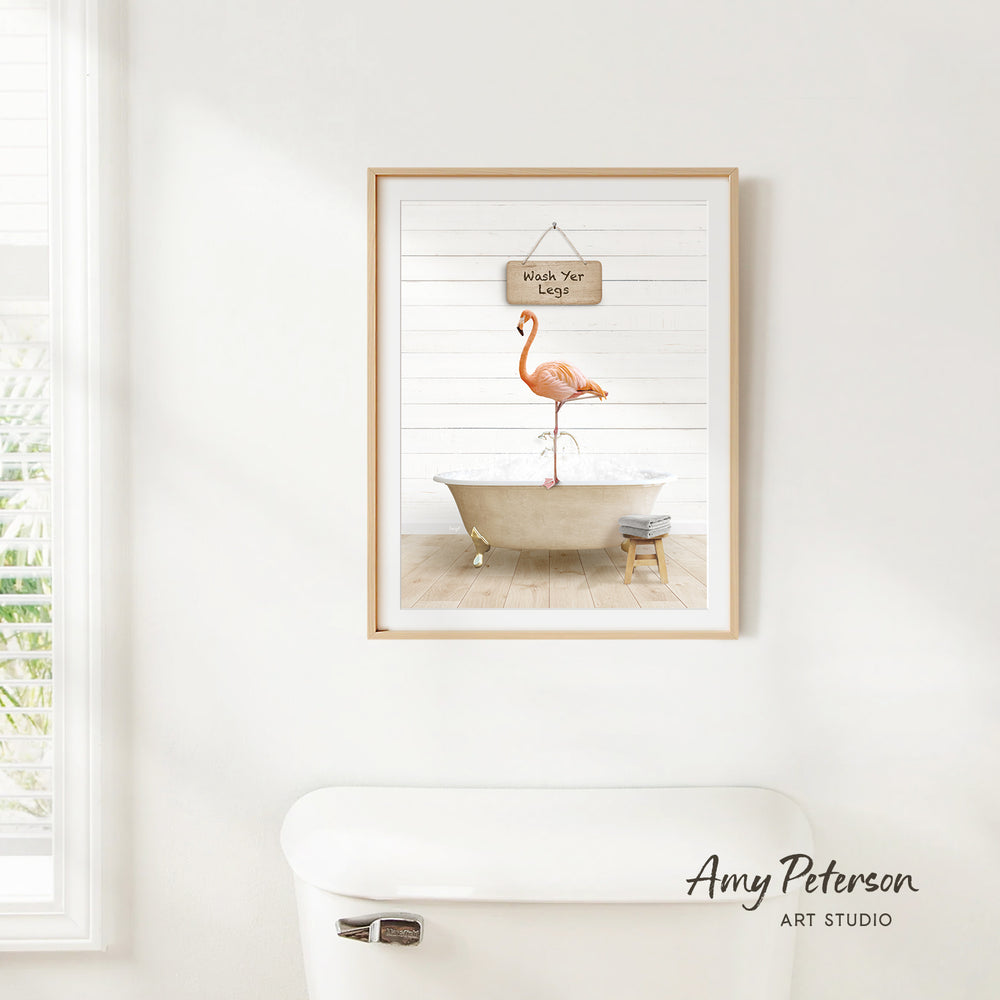 A framed print of a pink flamingo standing in a bathtub is hanging on a white wall above a white toilet.
