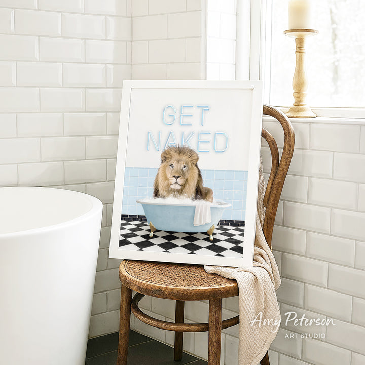 A framed image of a lion sitting in a bathtub with the text "GET NAKED" on it, placed on a wooden chair in a bathroom setting.