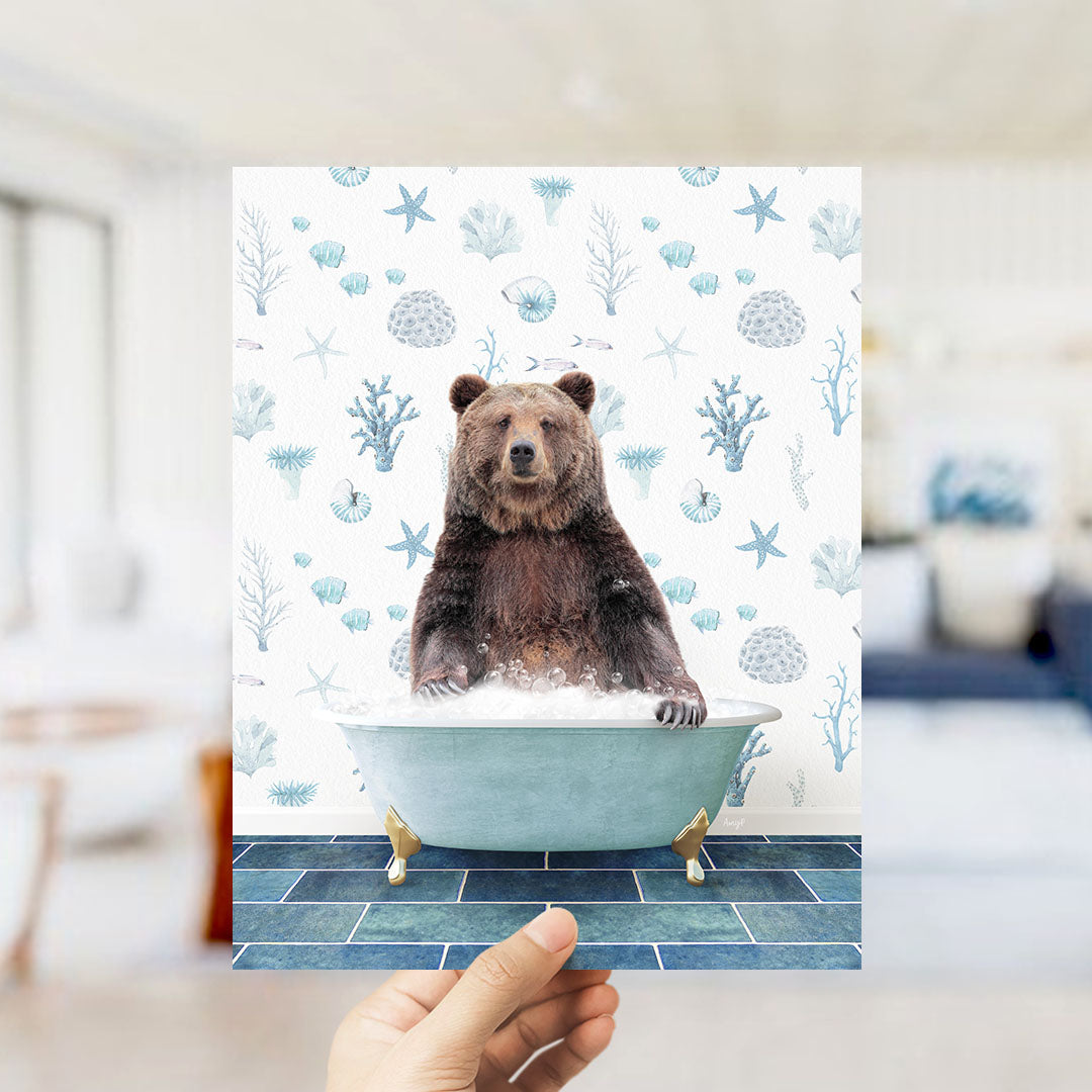 A hand is holding up a print featuring a brown bear sitting in a bathtub filled with bubbles, surrounded by a pattern of seashells and starfish.