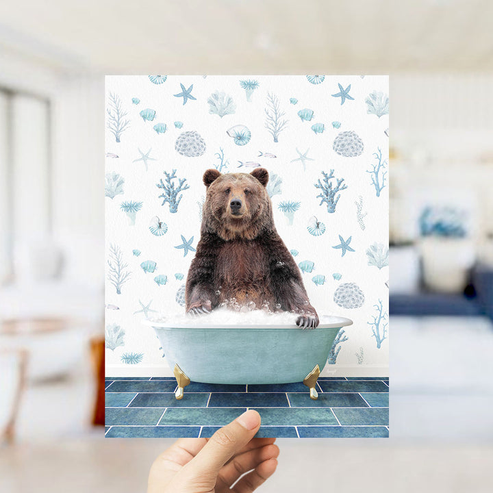 A hand is holding up a print featuring a brown bear sitting in a bathtub filled with bubbles, surrounded by a pattern of seashells and starfish.