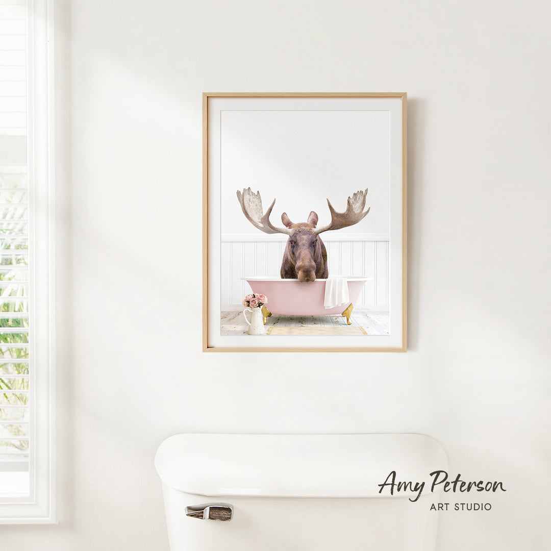 A framed image of a moose sitting in a pink bathtub, with a window and toilet visible in the background.