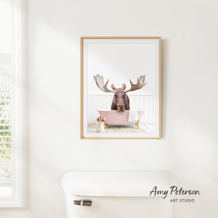 A framed image of a moose sitting in a pink bathtub, with a window and toilet visible in the background.