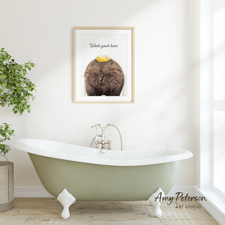 A framed print of a bear with a yellow object on its head hangs on a white wall above a green clawfoot bathtub in a bright, airy bathroom.