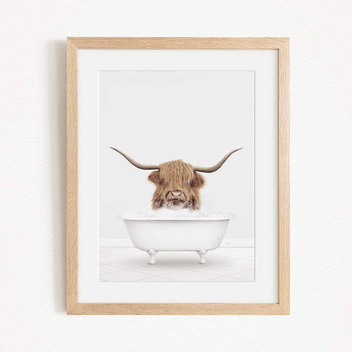 A framed image depicting a Highland cow with long horns sitting in a white bathtub filled with water.