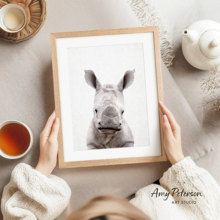 A person is holding a framed picture of a rhinoceros, with a teapot, a cup of tea, and a bowl of tea visible in the background.