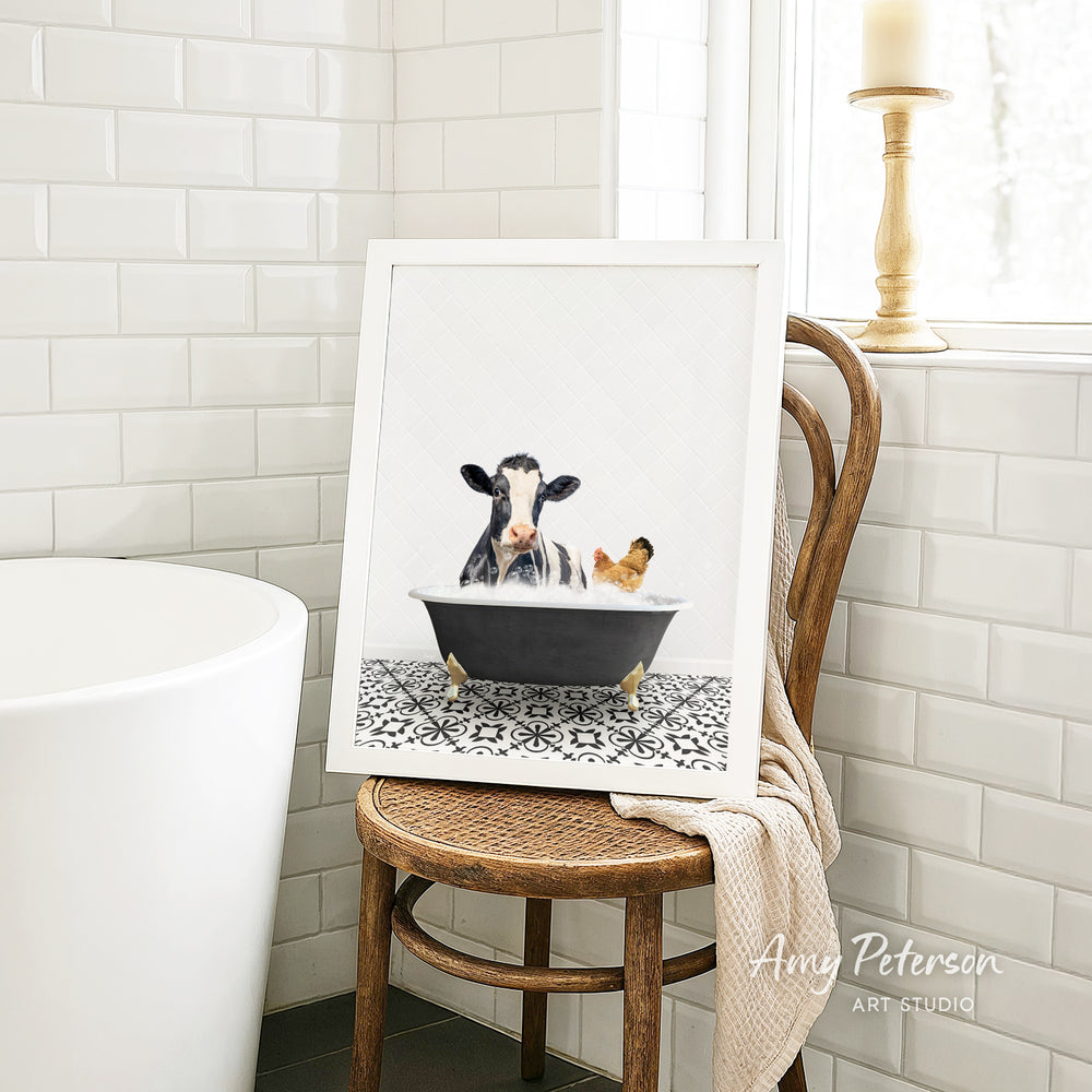 A framed image of a cow and a chicken sitting in a bathtub, with a towel draped over the back of a wooden chair in a bathroom setting.