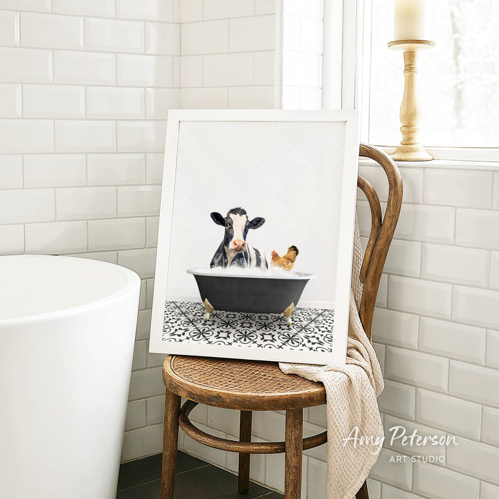 A framed image of a cow and a chicken sitting in a bathtub, with a towel draped over the back of a wooden chair in a bathroom setting.