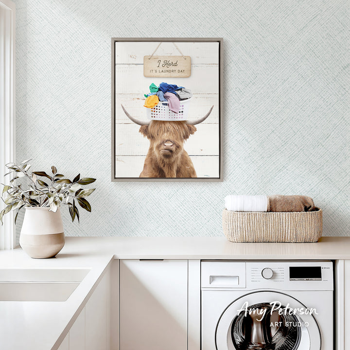 a framed picture of a Highland cow with a basket on its head, hanging on a wall above a white countertop with a plant and a basket of towels.