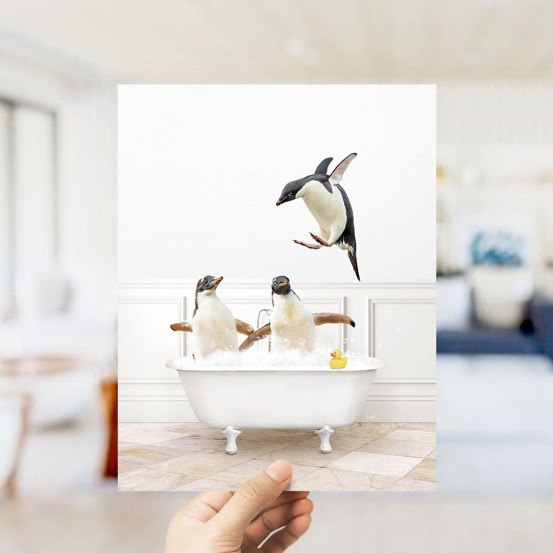 A hand holds up a framed photograph of two penguins sitting in a bathtub, with one penguin jumping out of the tub.