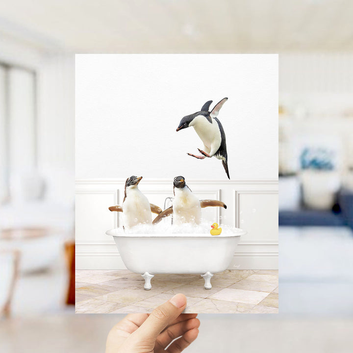 A hand holds up a framed photograph of two penguins sitting in a bathtub, with one penguin jumping out of the tub.