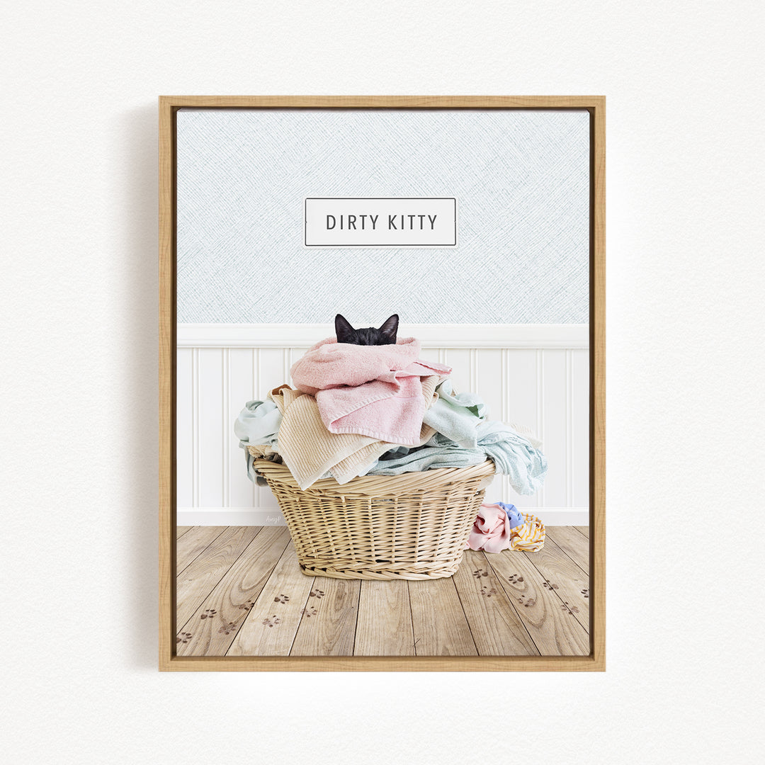 A framed image depicting a cat sitting in a basket filled with laundry, with a sign above the basket that says "Dirty Kitty".