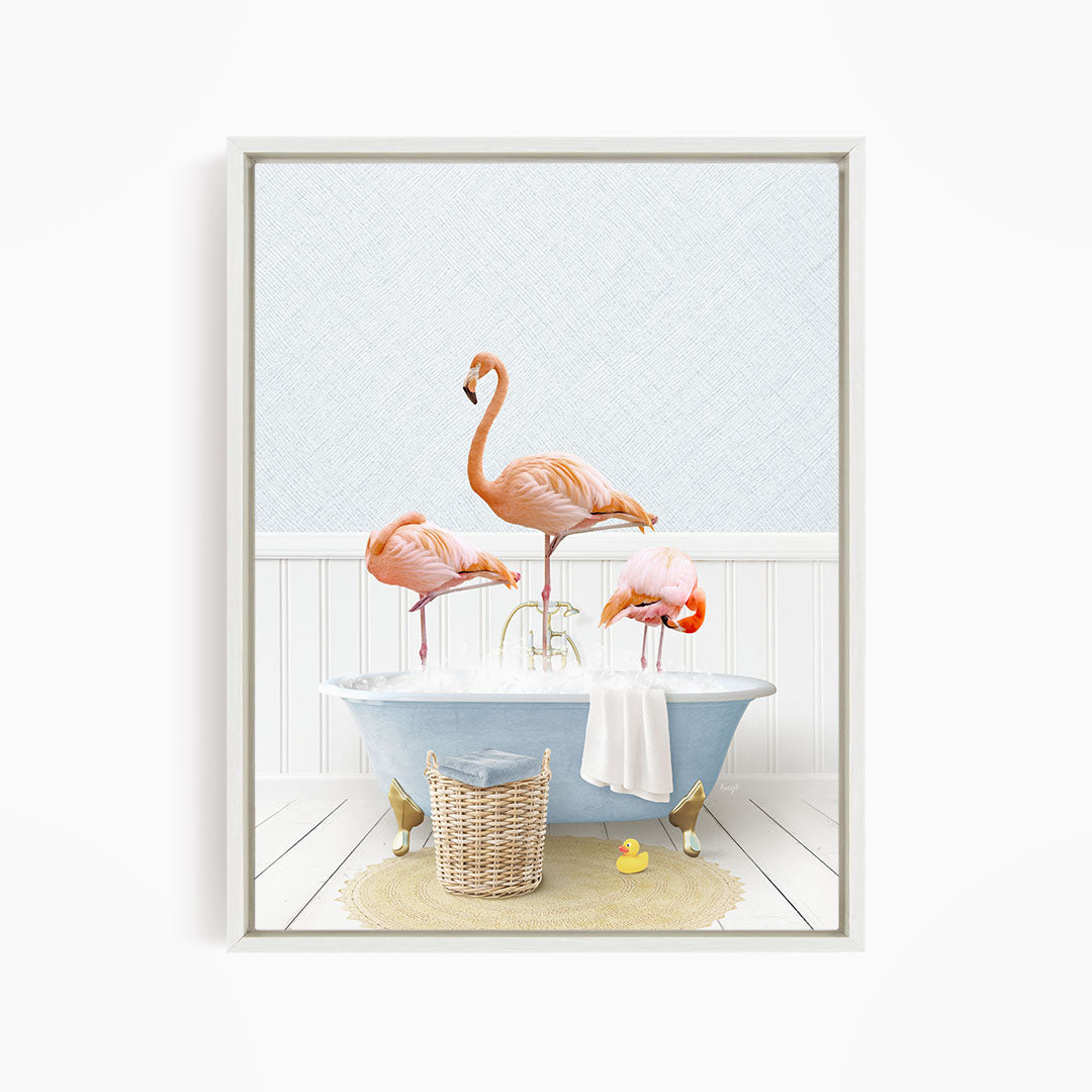 A framed image depicting three pink flamingos standing in a bathtub filled with water, with a basket of bath toys and a towel nearby.