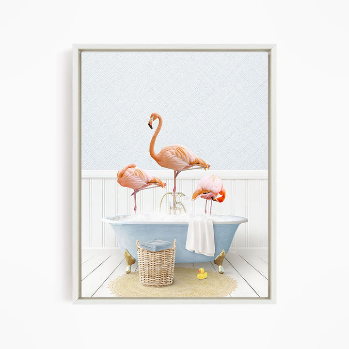 A framed image depicting three pink flamingos standing in a bathtub filled with water, with a basket of bath toys and a towel nearby.