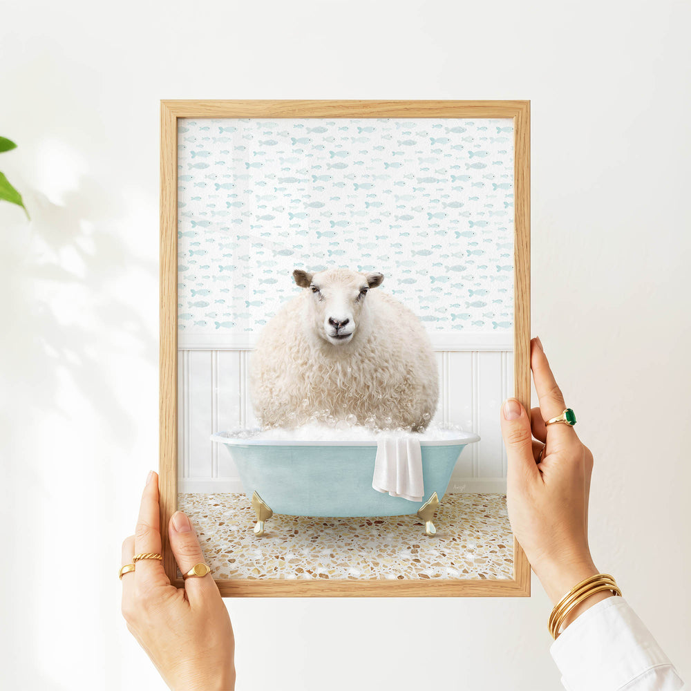 A person's hands are holding a framed image of a sheep sitting in a bathtub filled with bubbles, surrounded by a pattern of fish.