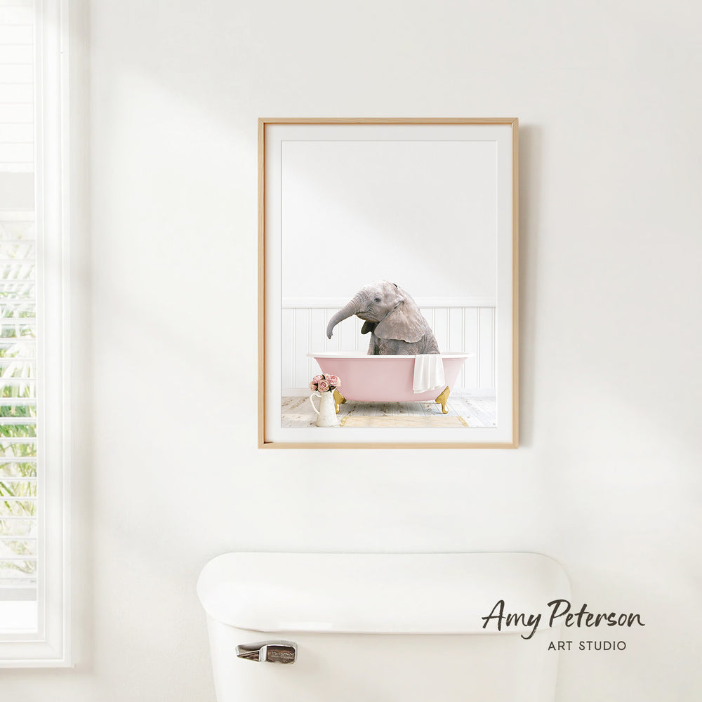 A framed photograph of a baby elephant sitting in a pink bathtub, with a white toilet and a window in the background.