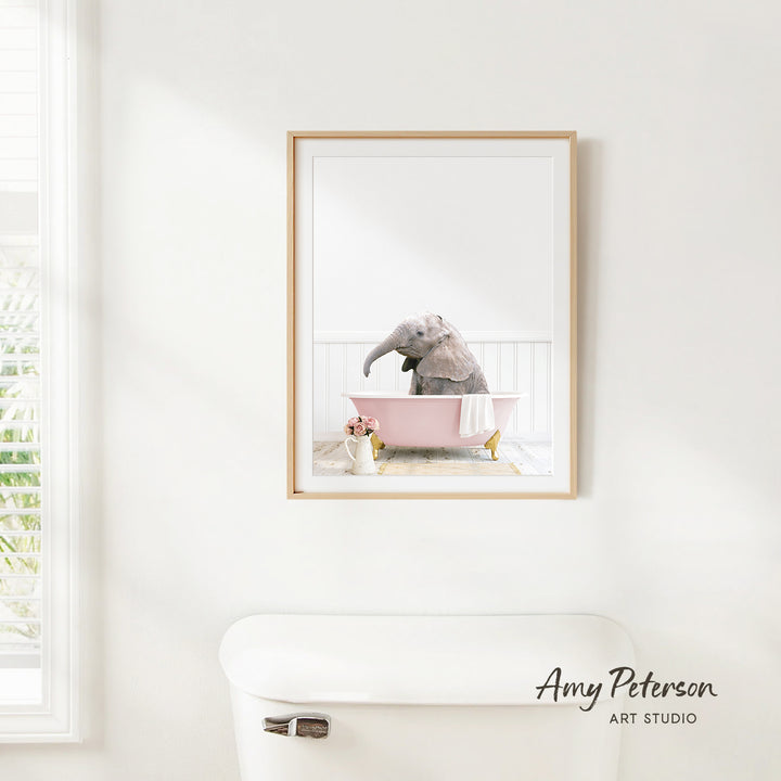 A framed photograph of a baby elephant sitting in a pink bathtub, with a white toilet and a window in the background.