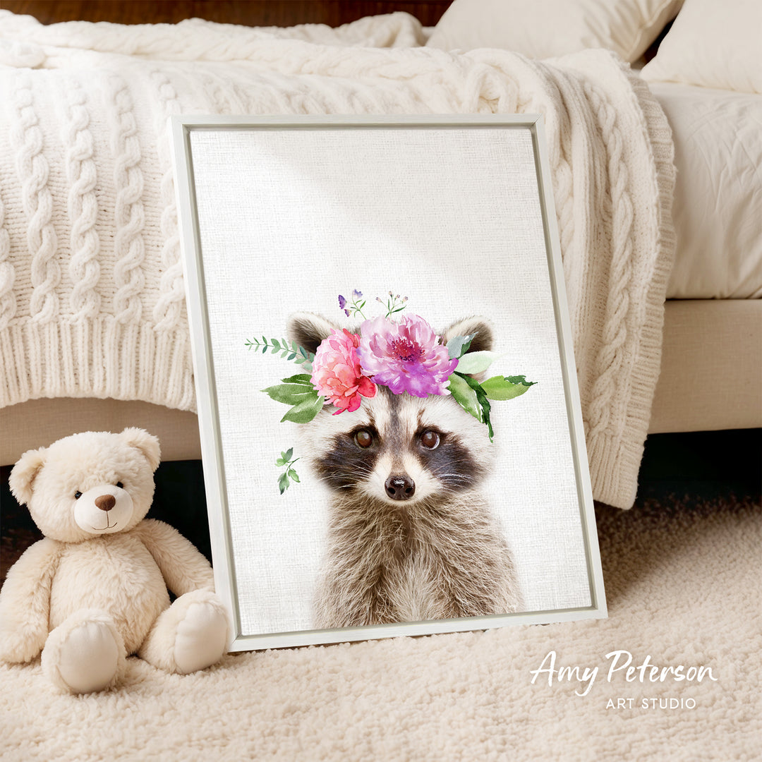 A framed print of a raccoon wearing a floral crown is displayed on a bed, accompanied by a teddy bear.