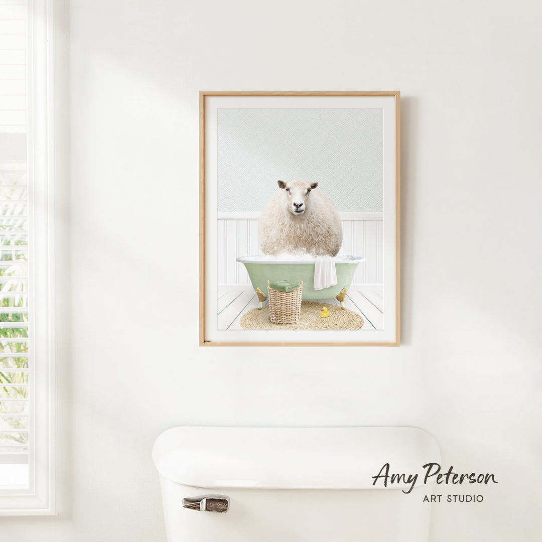 A framed image of a sheep sitting in a bathtub, surrounded by various bathroom items, such as a basket and a towel, is displayed on a white wall.