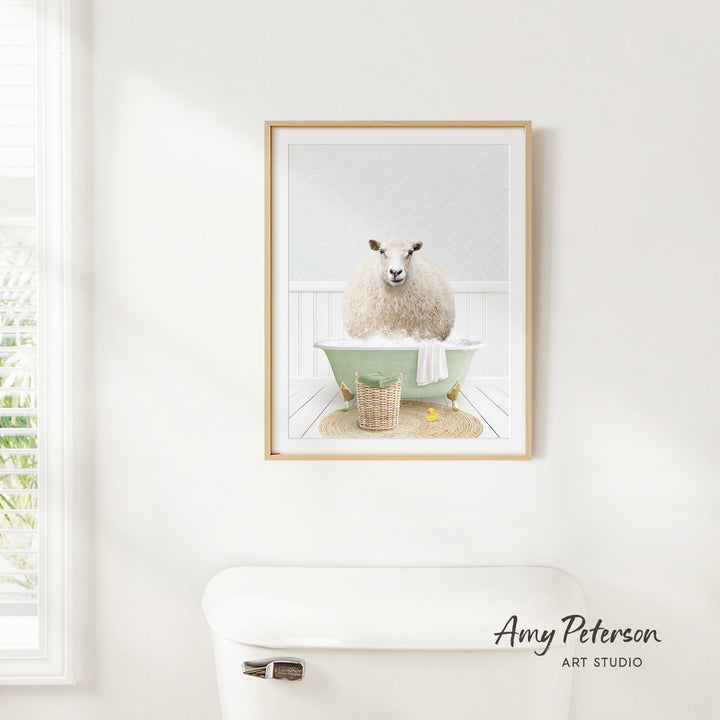 A framed image of a sheep sitting in a bathtub, surrounded by various bathroom items, such as a basket and a towel, is displayed on a white wall.