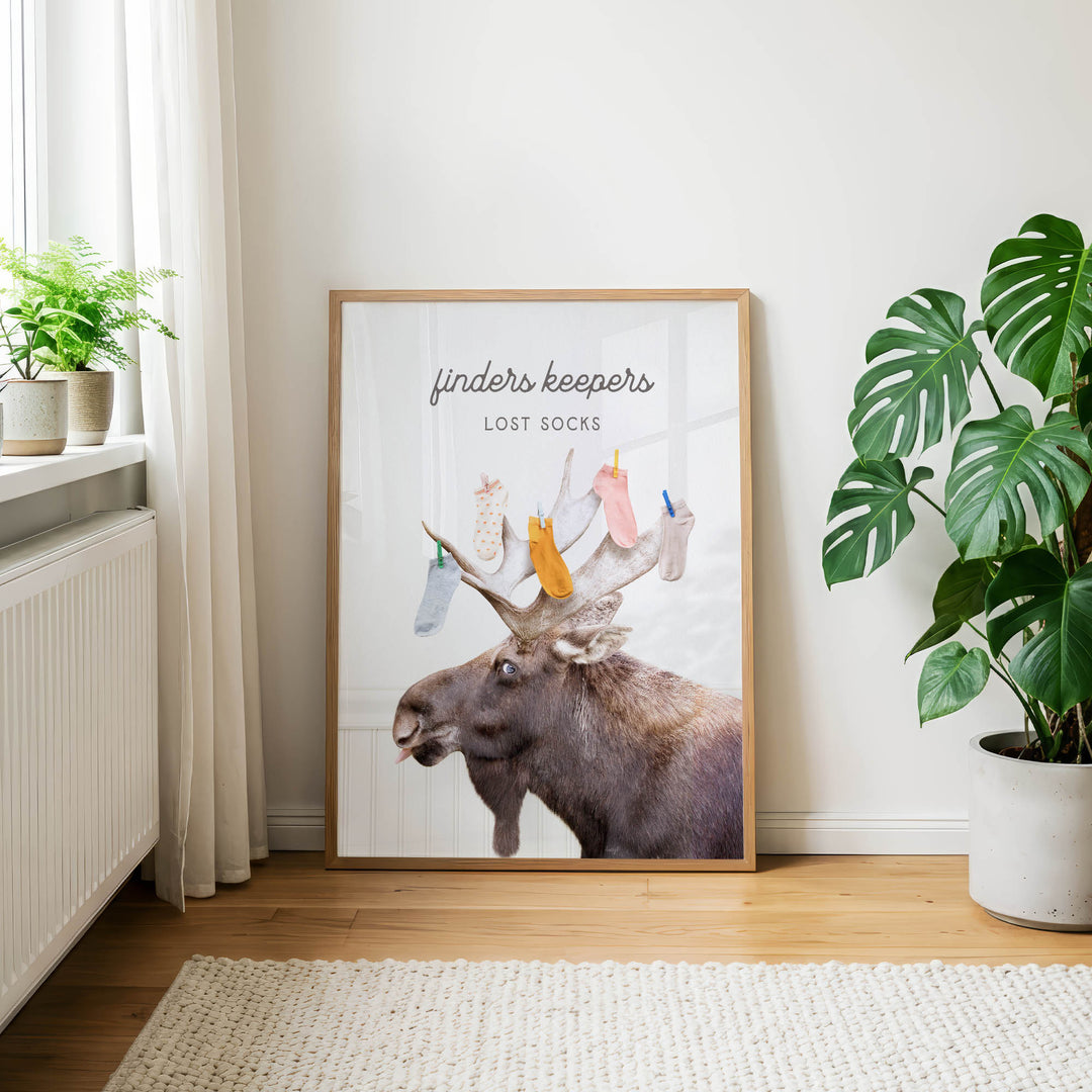 a framed poster of a moose with a humorous caption, "Finders keepers: Lost socks", placed on a wooden floor in a room with a window and potted plants.