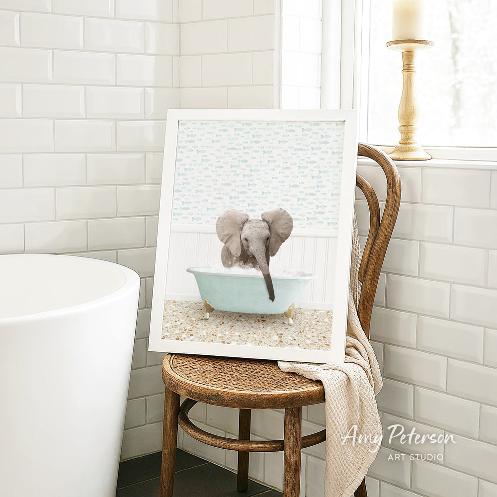 A framed image of a baby elephant sitting in a bathtub, surrounded by a light blue and white patterned backdrop, is displayed on a wooden chair in a bathroom setting.
