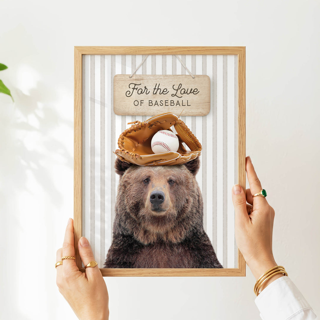 A person is holding a framed picture of a bear wearing a baseball cap and glove, with a sign above the bear that says "For the Love of Baseball".