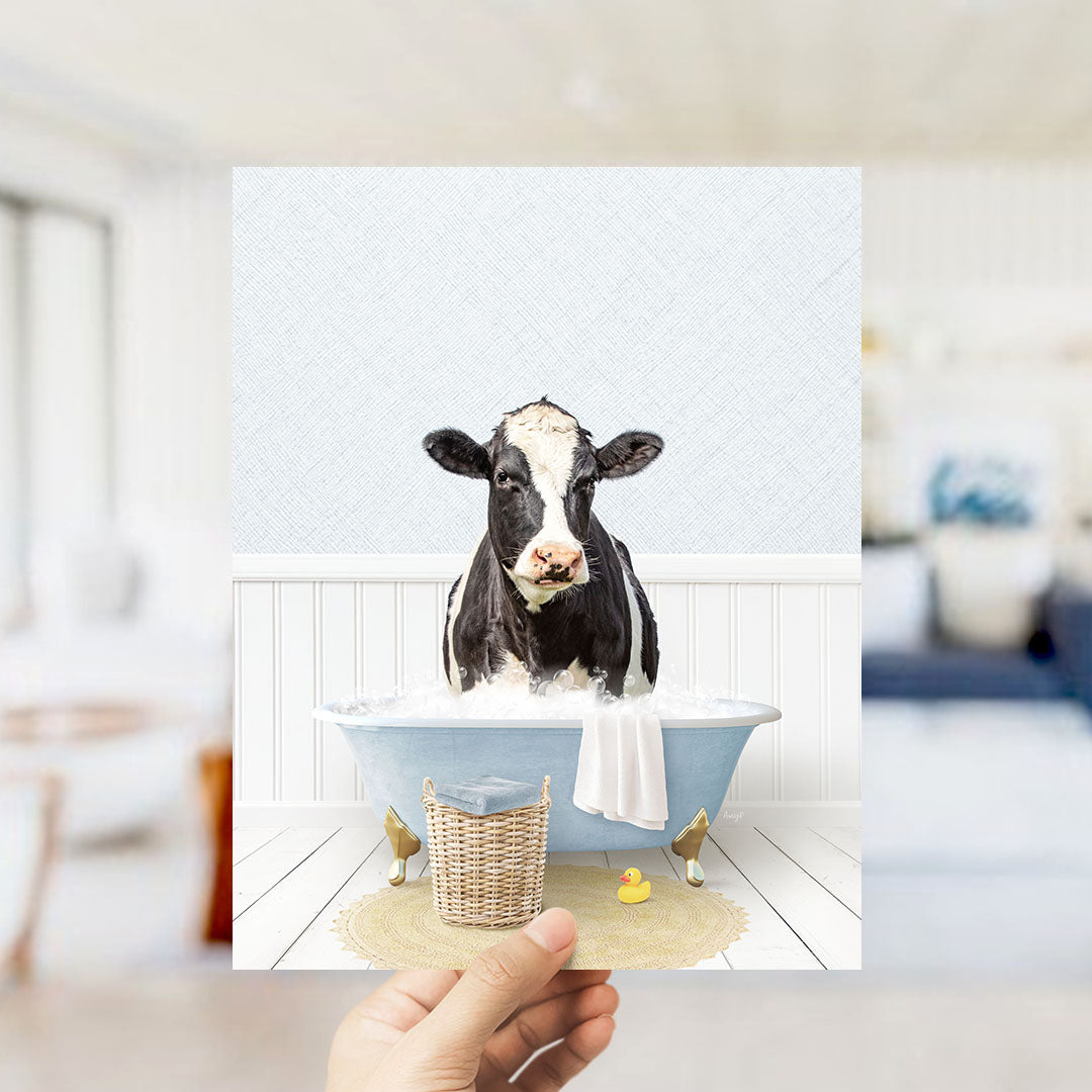 A black and white cow is sitting in a blue bathtub filled with water, surrounded by a yellow rubber duck and a white towel.