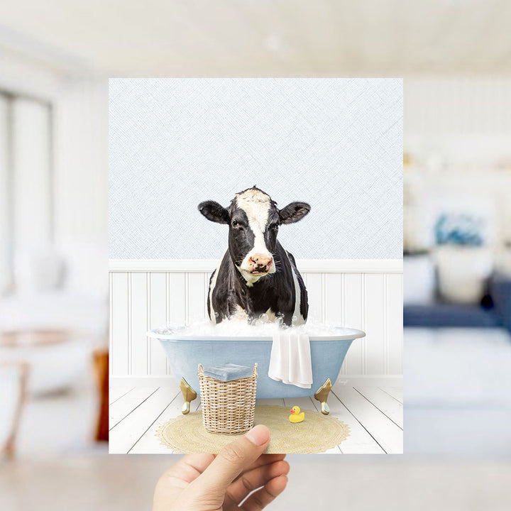 A black and white cow is sitting in a blue bathtub filled with water, surrounded by a yellow rubber duck and a white towel.